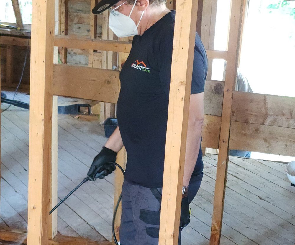 Close-up of an AtlantCare technician spraying antimicrobial sealer from a pump sprayer onto stripped framing